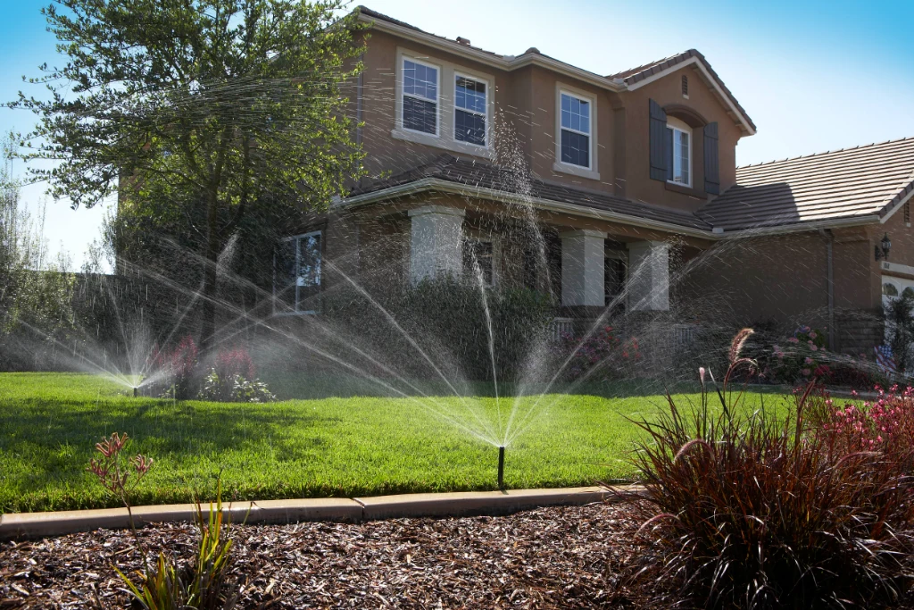 Sprinklers spraying across a green front lawn and mulched bed in front of a two-story home