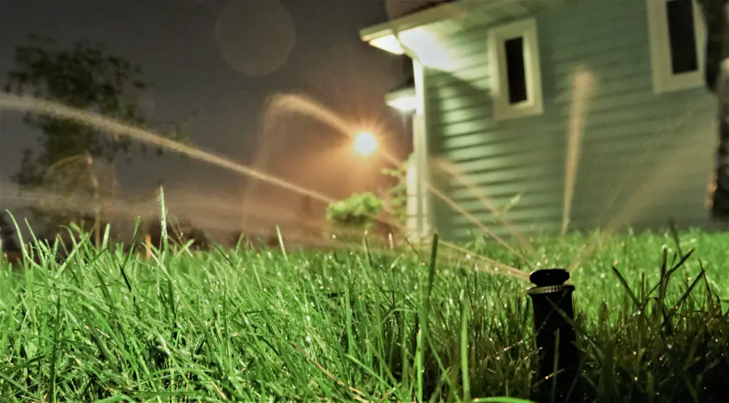 A sprinkler watering a lawn at night, with house lights illuminating the streams